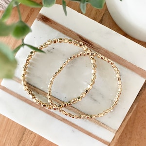 May include: Two gold beaded bracelets on a white marble surface with wood accents.