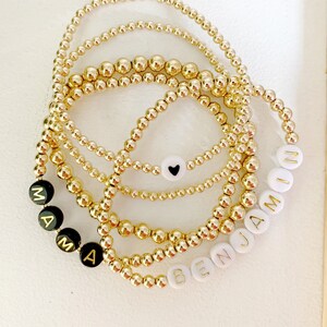 May include: Several gold beaded bracelets are displayed. Some bracelets have black beads with the word "MAMA" spelled out in gold letters. Other bracelets have white beads with the name "BENJAMIN" spelled out in gold letters. One bracelet has a white bead with a black heart.