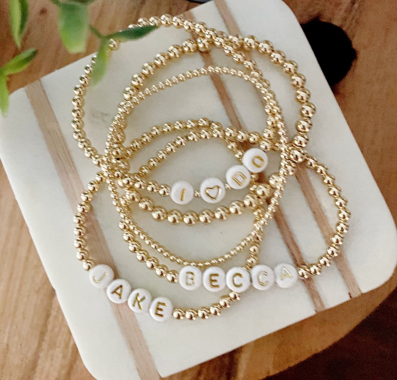 May include: Gold beaded bracelets with white letter beads spelling "JAKE", "BECCA", and "I DO" with a heart symbol. The bracelets are stacked on a white and tan marble-patterned surface.