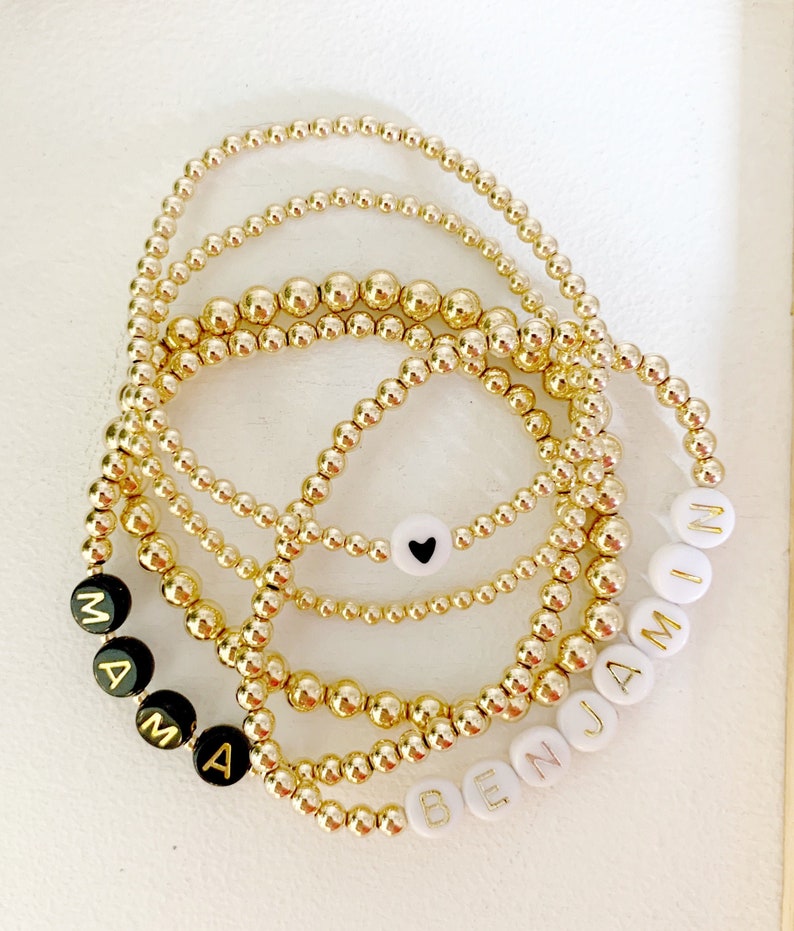May include: Several gold beaded bracelets are displayed. Some bracelets have black beads with the word "MAMA" spelled out in gold letters. Other bracelets have white beads with the name "BENJAMIN" spelled out in gold letters. One bracelet has a white bead with a black heart.