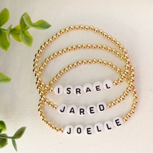 May include: Three gold bead bracelets with white letter beads spelling out the names "ISRAEL", "JARED", and "JOELLE".