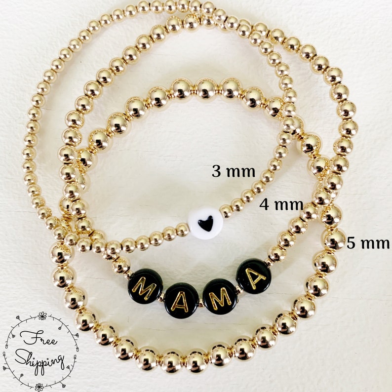 May include: Gold beaded bracelets in 3 mm, 4 mm, and 5 mm sizes. One bracelet features black beads spelling "MAMA", another has a white bead with a black heart. The bracelets are displayed on a white surface.