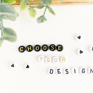 May include: White background with the words "CHOOSE YOUR DESIGN" spelled out with black and gold letter beads. Several white beads with black heart shapes are scattered around the text. Green leafy plant in the upper left corner.