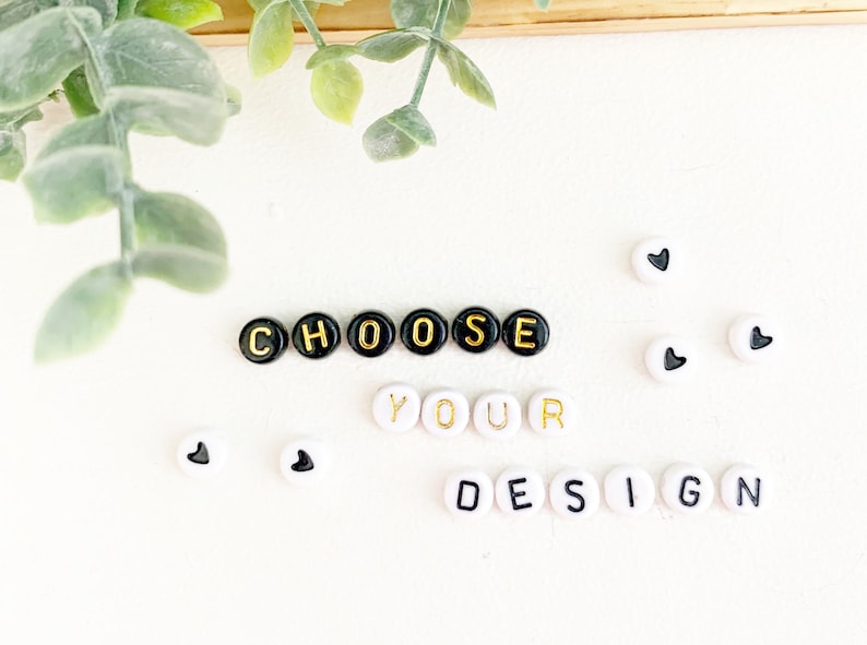 May include: White background with the words "CHOOSE YOUR DESIGN" spelled out with black and gold letter beads. Several white beads with black heart shapes are scattered around the text. Green leafy plant in the upper left corner.