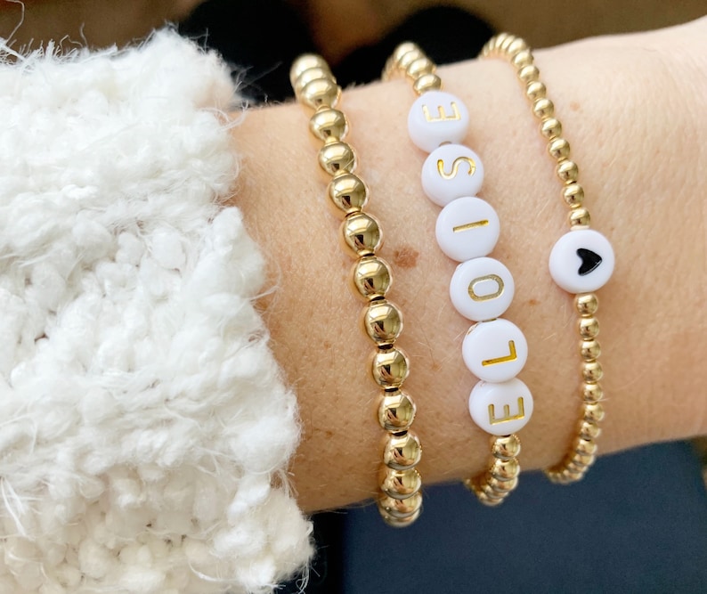 May include: Gold beaded bracelets, one spelling "LOVE" with gold letters on white beads, and another with a black heart on a white bead. The bracelets are worn on a wrist with a white, textured sleeve.