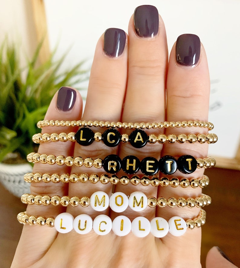 May include: Gold beaded bracelets with black and white letter beads spelling out names and the word "MOM". The bracelets are displayed on a hand with dark purple nail polish. The bracelets are a stylish accessory.