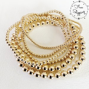 May include: A collection of gold beaded bracelets in varying sizes. The bracelets are arranged in a circular pattern, showcasing the different bead sizes and the overall shine of the gold. The image is set against a white background.