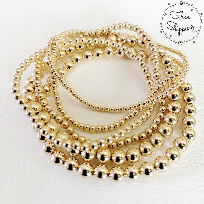 May include: A collection of gold beaded bracelets in varying sizes. The bracelets are arranged in a circular pattern, showcasing the different bead sizes and the overall shine of the gold. The image is set against a white background.
