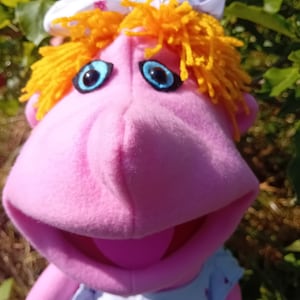 May include: A pink felt puppet featuring bright orange yarn hair, a white bow with a floral pattern, and large blue eyes. The puppet has a prominent nose, a wide open mouth, and wears a light blue dress with a small floral print on the bodice.