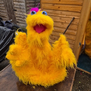 May include: A bright yellow fuzzy puppet featuring a wide open mouth with a pink tongue, two large blue eyes, and a tuft of vibrant pink hair on top. The puppet has two raised arms with three-fingered hands and is covered in long, shaggy faux fur.