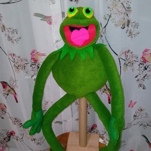 May include: A plush green frog puppet with large, round eyes, a pink tongue, and a wide open mouth. The puppet has long arms and legs, and is mounted on a wooden stand. The frog has a green collar.