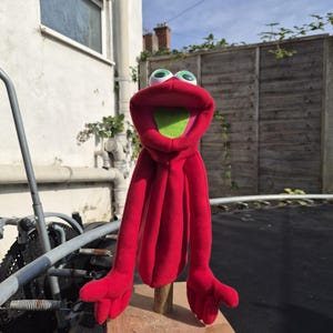 Puede incluir: Una marioneta de mano roja con la boca muy abierta y el interior de color verde brillante, grandes ojos blancos con pupilas verdes y largos brazos colgantes con manos de cuatro dedos, montada sobre un palo de madera y una base cuadrada.