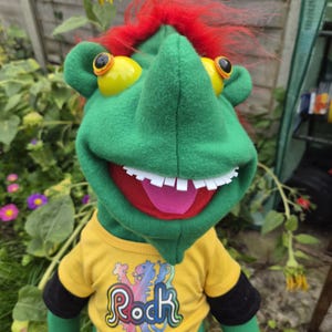 May include: A green puppet with bright red hair, large yellow eyes, and a wide mouth featuring white teeth and a pink tongue. The puppet wears a yellow short-sleeved shirt with black sleeves and the word Rock printed in colorful letters.