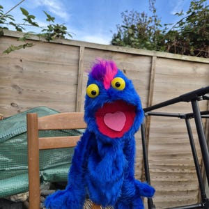May include: A blue fuzzy hand puppet with a bright pink mohawk, two large yellow eyes with black pupils, and an open mouth featuring a pink heart-shaped tongue. The puppet is mounted on a small wooden stand, showcasing its long shaggy fur.