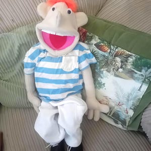 May include: A puppet with light skin, bright orange hair, and a wide open mouth with white teeth. It wears a short-sleeved shirt with horizontal light blue and white stripes, a small chest pocket, plain white trousers, and solid black shoes.