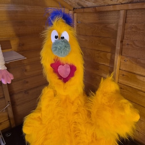 May include: A yellow fuzzy puppet featuring a blue tuft of hair on top, large white eyes, a round green nose, and an open mouth with a pink heart-shaped tongue. The puppet is covered in long, shaggy yellow faux fur and has two arms.