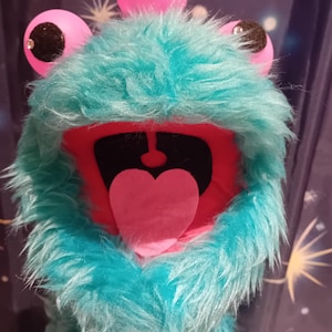May include: A plush monster puppet with three pink, googly eyes and a wide open mouth. The puppet has a turquoise fur body, a red mouth, and a pink heart-shaped tongue. The eyes have black pupils and are embellished with small rhinestones.