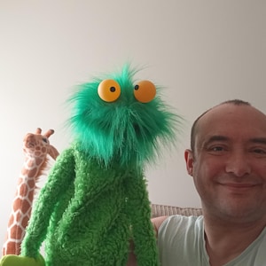 May include: A green fuzzy hand puppet with long shaggy fur on its head, two large round orange eyes with black pupils, and light green felt hands. The puppet has a textured green body and is positioned next to a plush giraffe toy.