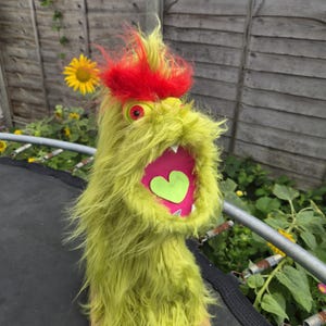 May include: A fuzzy lime green monster puppet with long shaggy fur, a bright red tuft of hair on top, and a single red eye. The puppet has a wide open mouth with a pink interior, white teeth, and a light green heart-shaped tongue inside.