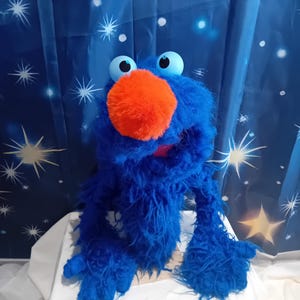 Op de afbeelding: Een blauwe pluchen pop met grote blauwe ogen en een oranje neus. De pop heeft een harige blauwe vacht en staat tegen een blauwe achtergrond met witte sterrenpatronen. De armen en handen van de pop zijn zichtbaar.