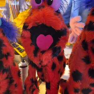 May include: A plush puppet featuring bright red fur with black spots, a shock of blue hair on top, large round purple eyes, and a prominent pink heart-shaped mouth. The creature has a soft, fuzzy texture and a wide, open expression.