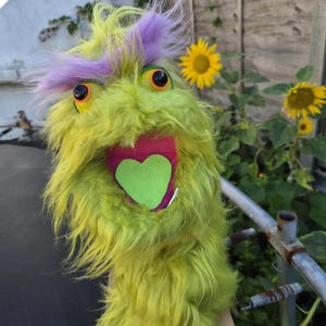 Op de afbeelding: Een pluizige, limoengroene monsterhandpop met lang, ruig imitatiebont, feloranje ogen en een plukje paars haar bovenop. De pop heeft een open mond met een roze binnenkant en een opvallende, lichtgroene tong in de vorm van een hartje.