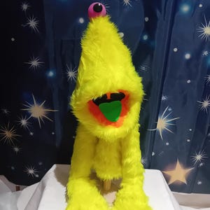 Op de afbeelding: Een gele pluchen pop met een kegelvormig hoofd, een roze oog en een brede mond die een rood interieur en een groene tong laat zien. De pop heeft lange, harige armen en voeten. Het wordt gepresenteerd op een wit oppervlak tegen een sterrenblauwe achtergrond.