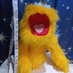 Könnte beinhalten: Eine skurrile, gelbe, pelzige Marionette mit einem großen roten Mund und einem rosa Herzen. Die Marionette hat zwei blaue Augen und lange, zottelige Arme. Die Marionette ist etwa 46 cm hoch.