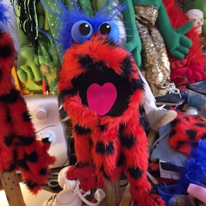 May include: A hand puppet covered in bright red and black spotted faux fur. It features large blue eyes with white pupils, a tuft of blue hair on top, and a wide open mouth with a prominent pink heart-shaped tongue inside the dark interior.