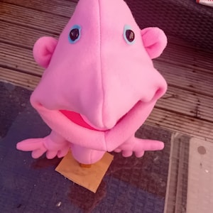 May include: A pink fleece puppet featuring a pointed head, two small ears, and a wide open mouth with a bright pink interior. The puppet has two dark circular eyes surrounded by light blue felt patches and two small arms with four fingers each.