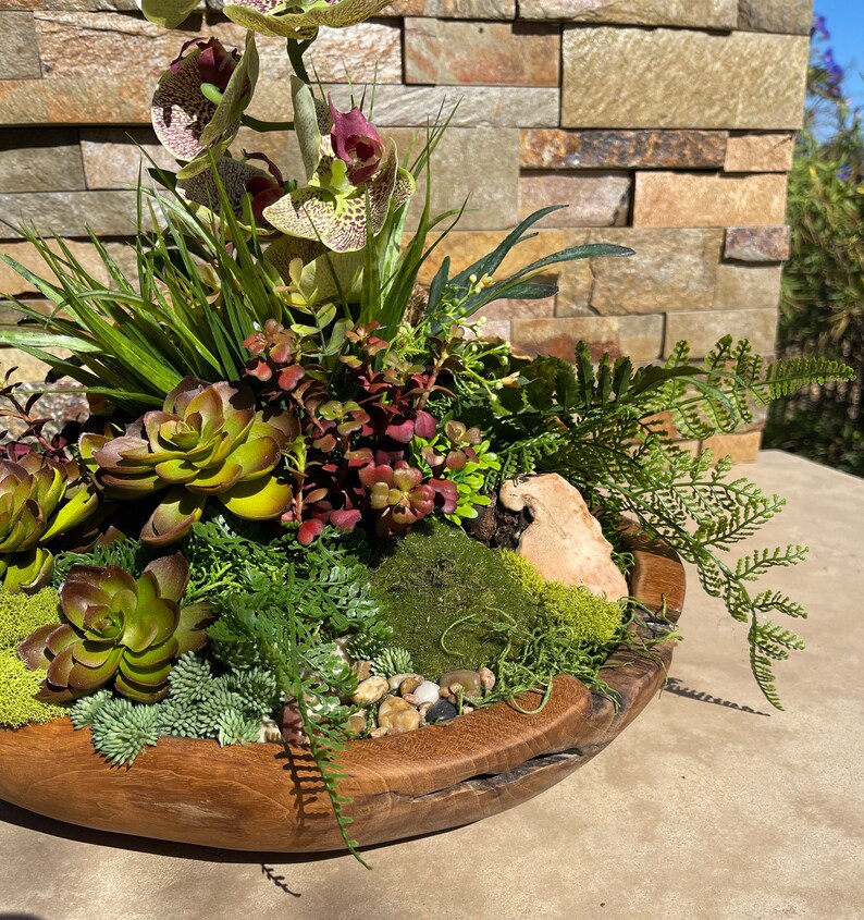 May include: A wooden bowl filled with a succulent arrangement featuring green and brown succulents, moss, and a fern. The bowl is decorated with small white stones.