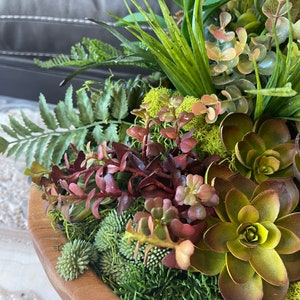 May include: A wooden bowl filled with a variety of green and brown succulents, ferns, and moss. The bowl is made of natural wood and has a rustic look.