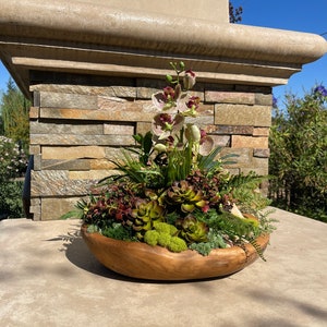 May include: A wooden bowl filled with a succulent arrangement. The arrangement features a variety of succulents, including green, brown, and red varieties. The bowl is sitting on a light brown surface.