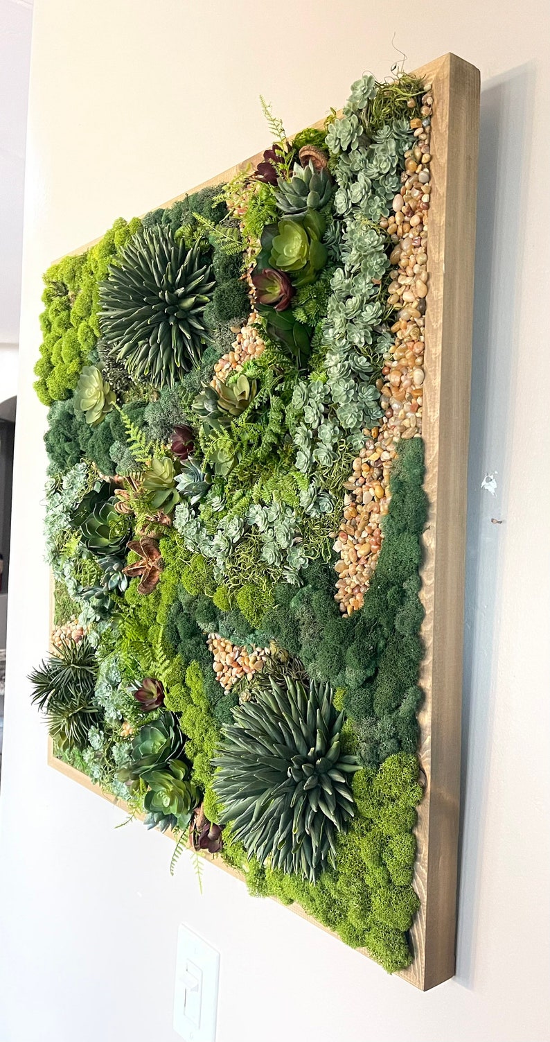 Puede incluir: Una pieza de arte de pared enmarcada que presenta una variedad de suculentas verdes y musgo, con peque&ntilde;as piedras beige intercaladas. El marco est&aacute; hecho de madera marr&oacute;n claro.