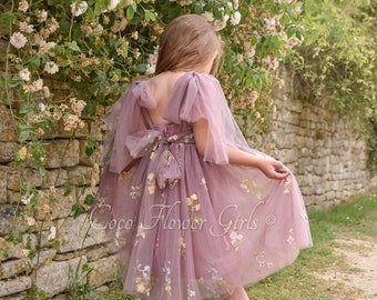 May include: A mauve tulle dress with embroidered flowers and a bow at the back. The dress has a scoop neckline and short sleeves. The dress is worn by a child standing in a garden.