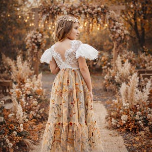 May include: A floor-length dress featuring a white lace bodice with short ruffled sleeves and a deep V-back. The skirt is made of sheer beige fabric adorned with intricate, multi-colored floral embroidery in shades of orange, brown, and green.