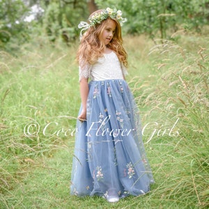 May include: A young girl wearing a light blue tulle dress with floral embroidery and a white lace bodice. She is wearing a flower crown and standing in a field of tall grass. The dress has a full skirt and is perfect for a special occasion.