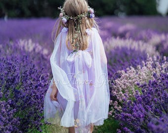 May include: A young girl wearing a white dress with a floral pattern and a white sheer cape walks through a field of purple lavender flowers. She is wearing a flower crown with pink and white flowers.