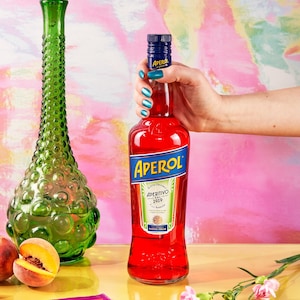 Könnte beinhalten: Ein lebendiges Bild mit einer Flasche Aperol, einer grünen Glasvase und einem Pfirsich. Die Aperol-Flasche ist rot mit einem blauen Etikett. Die Vase ist hoch und grün mit einer strukturierten Oberfläche. Der Hintergrund ist rosa und lila.