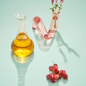 Może przedstawiać: Martwa natura z przezroczystą szklaną karafką wypełnioną bursztynowym płynem, różowym szklanym wazonem z fioletowymi kwiatami i kiścią czerwonych winogron. Obiekty rzucają cienie na jasnoniebieską powierzchnię.