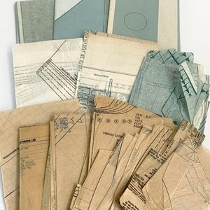 May include: A collection of translucent sewing pattern pieces made of thin, crinkled paper. The stack includes various tan and light blue sheets featuring printed black lines, markings, and text for garment construction and cutting guides.