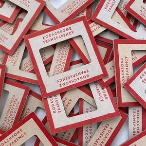 May include: A collection of vintage Kodachrome transparency mounts. Each mount is rectangular with a red border and a central square opening. The text "KODACHROME TRANSPARENCY" is printed in red on each mount, along with "PROCESSED BY KODAK".