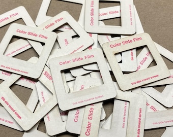 May include: A collection of square white cardboard color slide film mounts scattered in a pile. Each mount features a central square cutout and is printed with the text Color Slide Film and the instruction this side towards screen in pink ink.