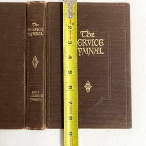 May include: Three brown hardcover books with gold lettering and decorative elements. The book on the right is measured with a tape measure, showing a height of approximately 8 inches. The text on the books reads "The SERVICE HYMNAL" and "HOPE PUBLISHING COMPANY."