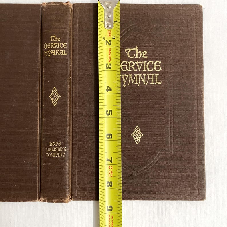 May include: Three brown hardcover books with gold lettering and decorative elements. The book on the right is measured with a tape measure, showing a height of approximately 8 inches. The text on the books reads "The SERVICE HYMNAL" and "HOPE PUBLISHING COMPANY."