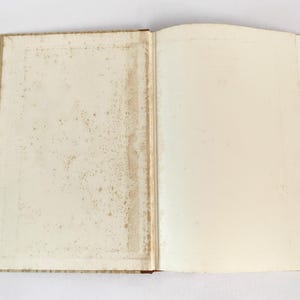 May include: An open book with aged, off-white pages. The left page shows signs of age with brown staining and spotting, while the right page is mostly clean. The book's spine is visible, suggesting a vintage or antique item.