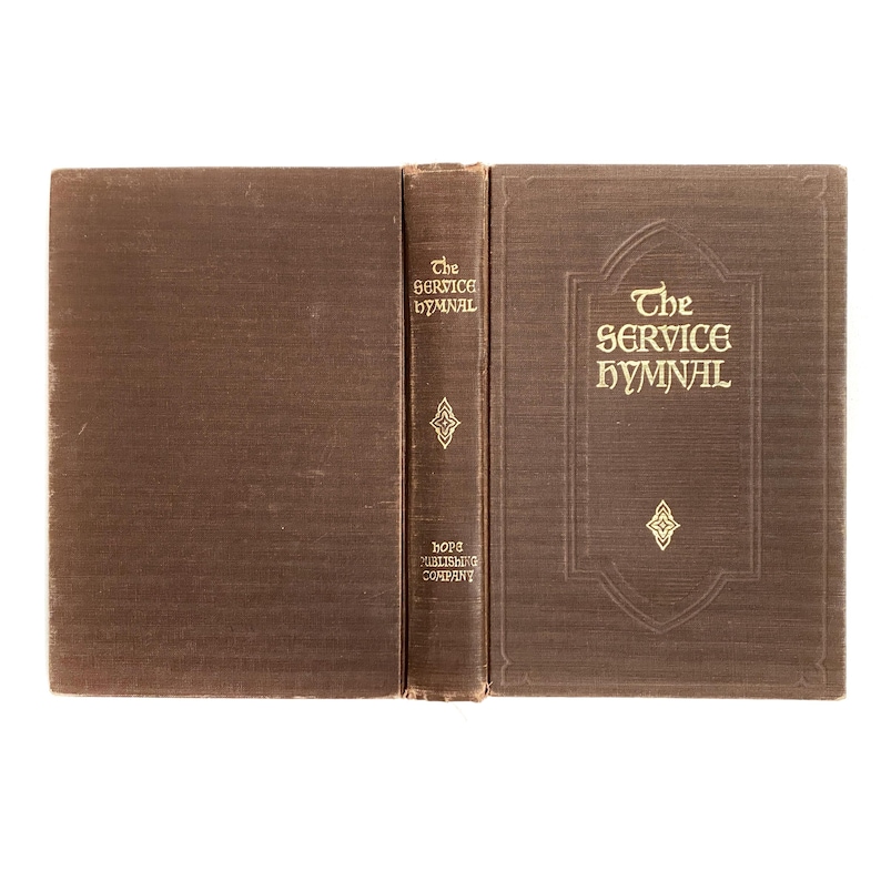 May include: An open brown book titled "The Service Hymnal" with gold lettering. The book is displayed open, revealing the spine and cover. The spine reads "The Service Hymnal" and "Hope Publishing Company".