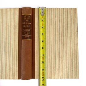 May include: A vintage book titled "Three Plays of Henrik Ibsen" with a brown spine and cover. The book is open, revealing pages with a striped pattern in beige and light green. A yellow tape measure shows the book is approximately 10 inches tall.