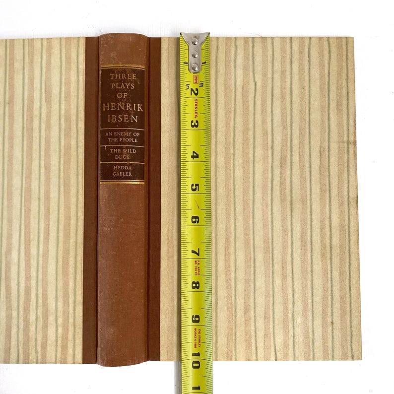 May include: A vintage book titled "Three Plays of Henrik Ibsen" with a brown spine and cover. The book is open, revealing pages with a striped pattern in beige and light green. A yellow tape measure shows the book is approximately 10 inches tall.