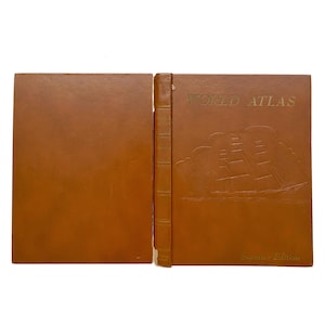 May include: An open, vintage world atlas with a brown leather cover. The cover features embossed text reading "WORLD ATLAS" and a ship illustration. The words "Superior Edition" are printed at the bottom. The book is open, revealing the inside pages.